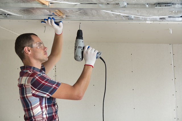 Young man in goggles fixing drywall suspended ceiling to metal frame using electrical screwdriver on ceiling insulated with shiny aluminum foil. Renovation, construction, do it yourself concept.